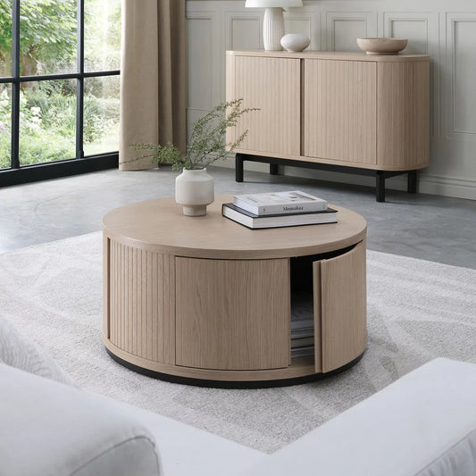 Bentley Designs Vega Scandi light oak round coffee table with natural grain top and dark peppercorn pedestal base in a bright living room setting.