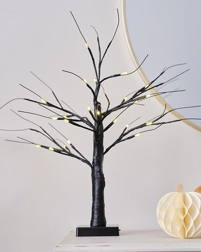 A small, matte black decorative tree with glowing warm white LED lights on the branches, sitting illuminated on a modern wooden entryway table.
