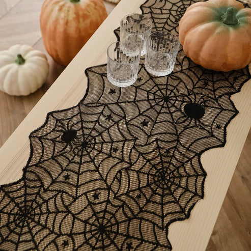 Top-down view of a black spider web lace table runner laid across a light wooden dining table with rustic Halloween decorations.