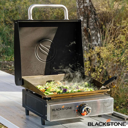 Angle view of the black Blackstone 17 inch tabletop gas griddle with open hood standing on an outdoor wooden table.