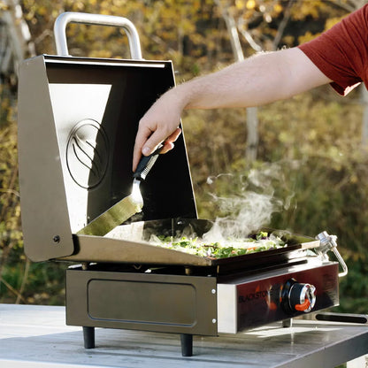 Angle view of the black Blackstone 17 inch tabletop gas griddle with open hood standing on an outdoor wooden table.