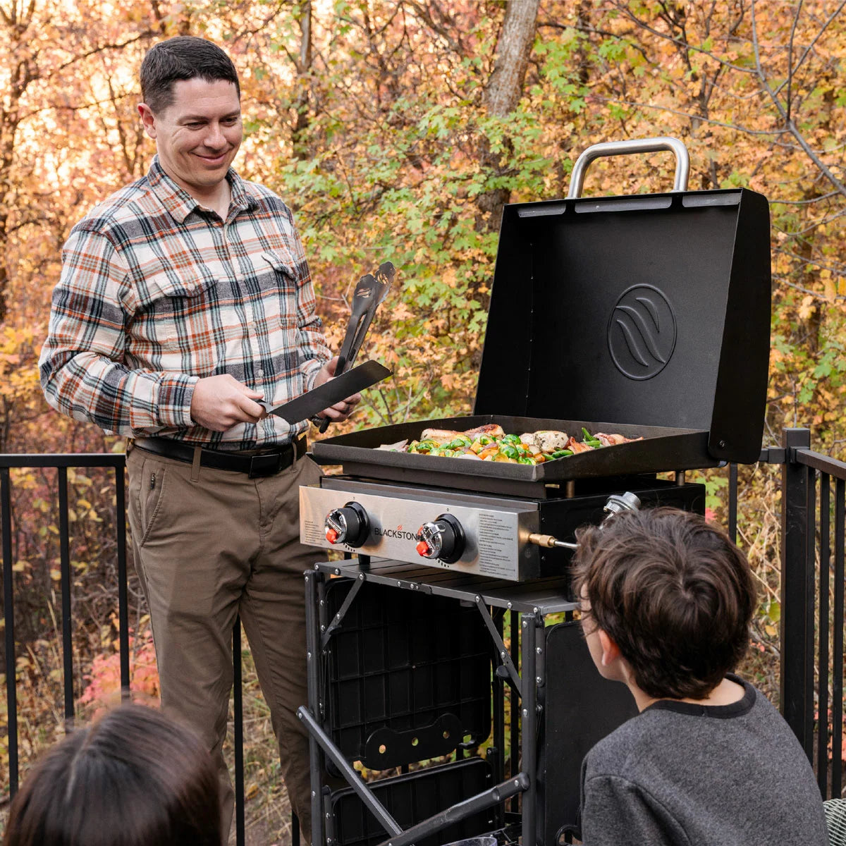The Blackstone 22" Table Top Gas Griddle with Hood displayed outdoors, showcasing its stainless steel burners, flat cooking plate, and integrated protective lid.