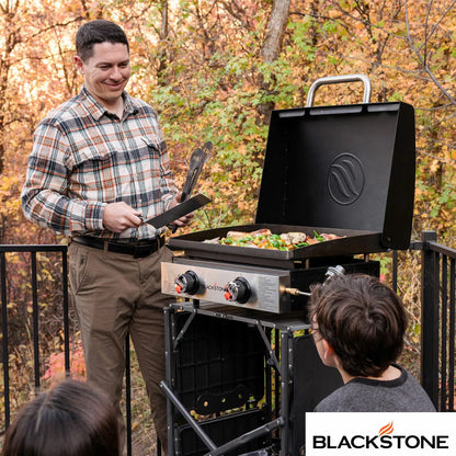 Angle view of the black Blackstone 22 inch tabletop gas griddle showing the open hood, dual burner dials, and flat steel cooking plate resting on a wooden bench.