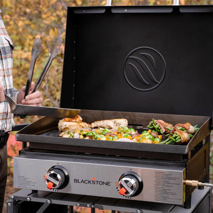 Angle view of the black Blackstone 22 inch tabletop gas griddle showing the open hood, dual burner dials, and flat steel cooking plate resting on a wooden bench.