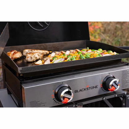 Angle view of the black Blackstone 22 inch tabletop gas griddle showing the open hood, dual burner dials, and flat steel cooking plate resting on a wooden bench.