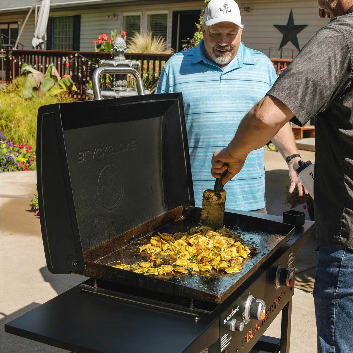 Front three-quarter view of the black Blackstone 28 inch gas griddle with hood open, showing the raw steel flat cooktop and foldable side shelves on a stone patio.