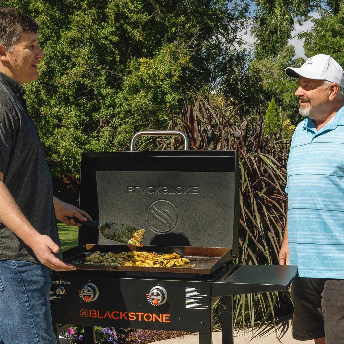 Front three-quarter view of the black Blackstone 28 inch gas griddle with hood open, showing the raw steel flat cooktop and foldable side shelves on a stone patio.