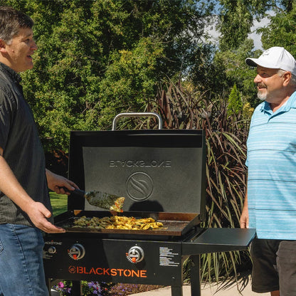 Front three-quarter view of the black Blackstone 28 inch gas griddle with hood open, showing the raw steel flat cooktop and foldable side shelves on a stone patio.