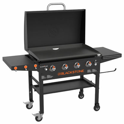 Angle view of the large black 4 burner Blackstone 36 inch gas griddle with hood raised, showing the massive steel cooktop, folded side shelves, and caster wheels on a patio.