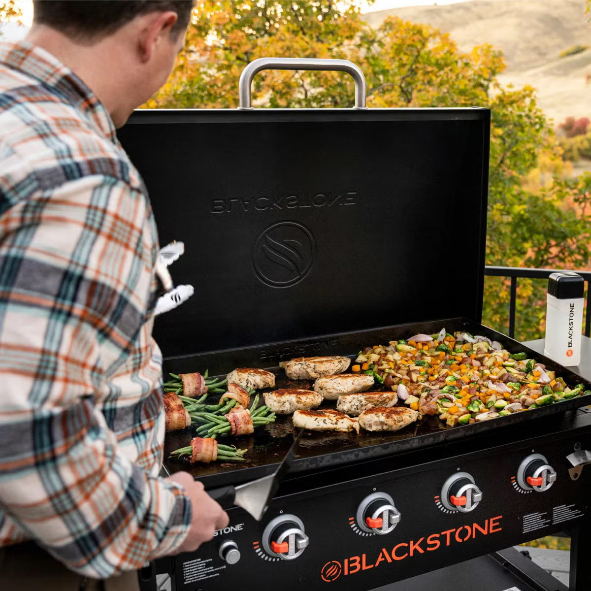 Angle view of the large black 4 burner Blackstone 36 inch gas griddle with hood raised, showing the massive steel cooktop, folded side shelves, and caster wheels on a patio.