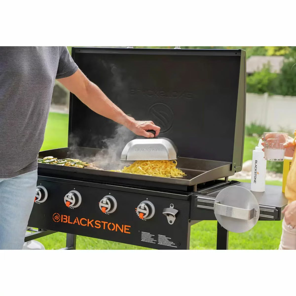 Angle view of the large black 4 burner Blackstone 36 inch gas griddle with hood raised, showing the massive steel cooktop, folded side shelves, and caster wheels on a patio.