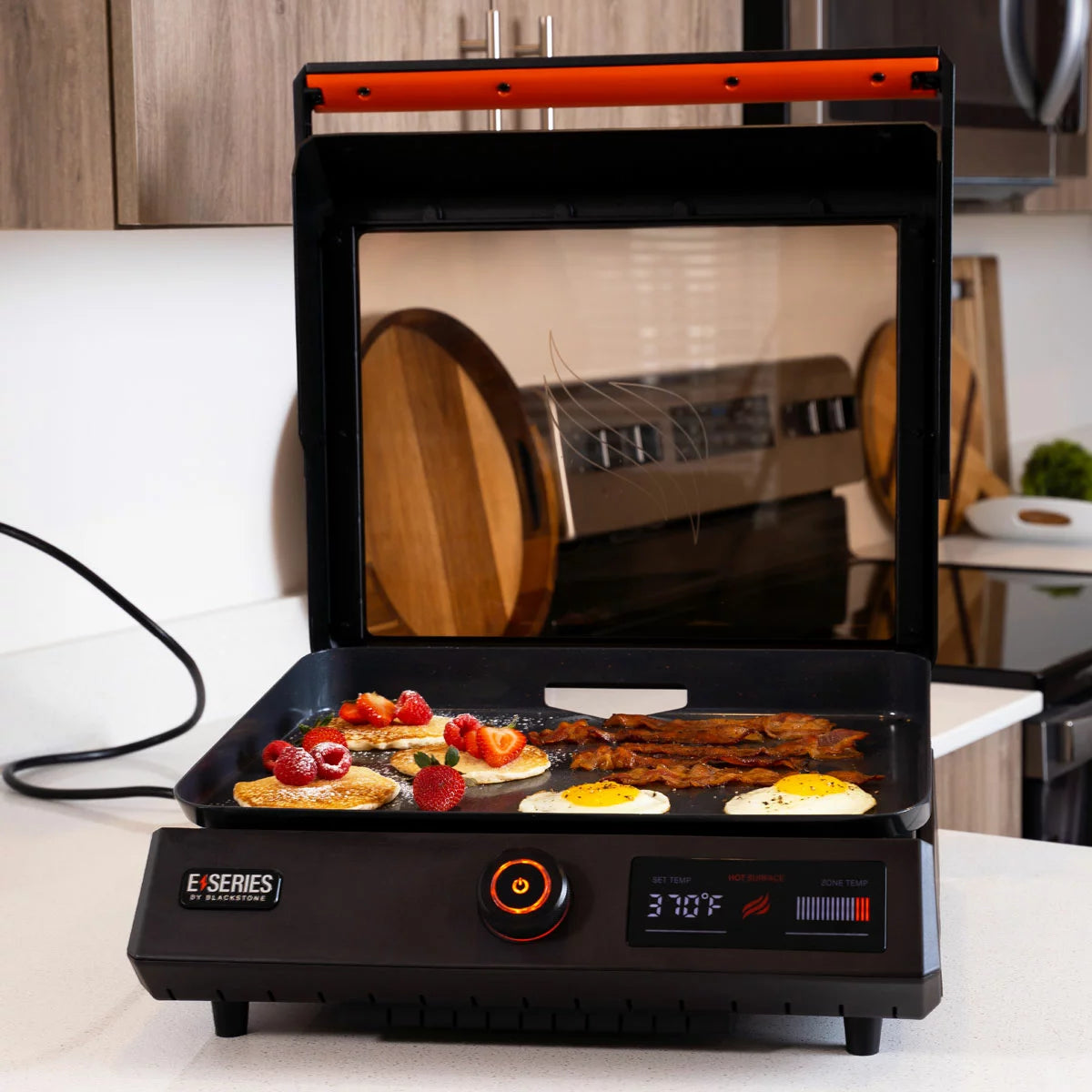 A Blackstone E-Series 17" electric griddle in a modern kitchen setting, showing the sleek glass hood, digital LCD control panel, and large non-stick ceramic cooking surface.