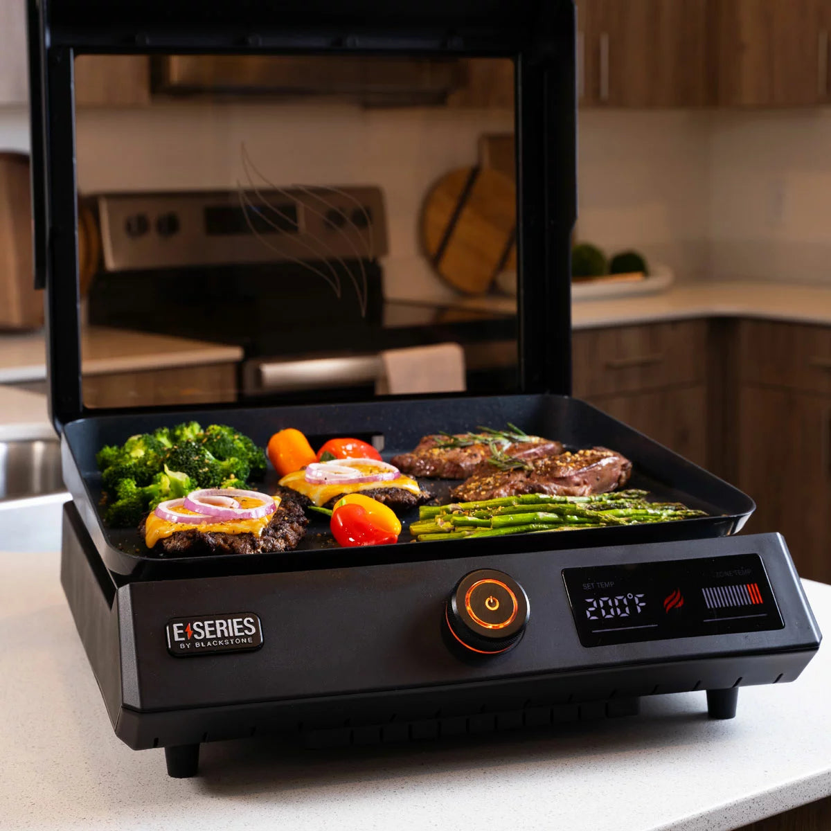 A Blackstone E-Series 17" electric griddle in a modern kitchen setting, showing the sleek glass hood, digital LCD control panel, and large non-stick ceramic cooking surface.