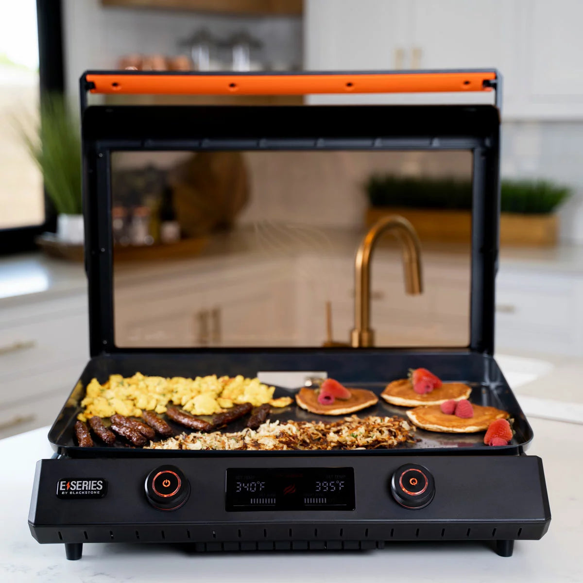 The Blackstone E-Series 22" Electric Griddle with its sleek glass hood and digital control panel, set up on a modern kitchen counter with cooked food ready to serve.