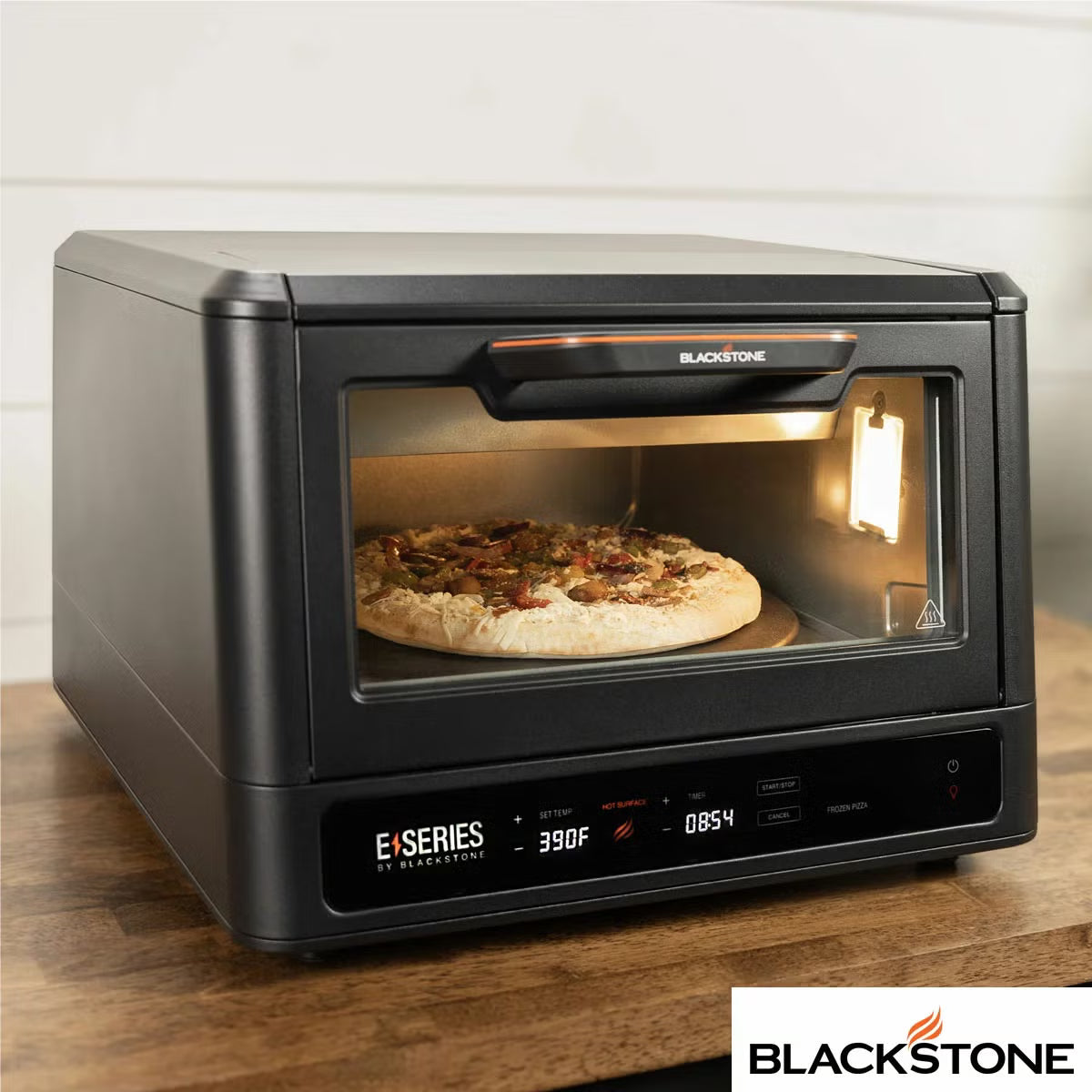 Front view of the black Blackstone E-Series electric pizza and air fryer oven sitting on a kitchen counter displaying its touchscreen control panel and open glass door with a 14 inch rotating deck inside.