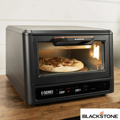 Front view of the black Blackstone E-Series electric pizza and air fryer oven sitting on a kitchen counter displaying its touchscreen control panel and open glass door with a 14 inch rotating deck inside.
