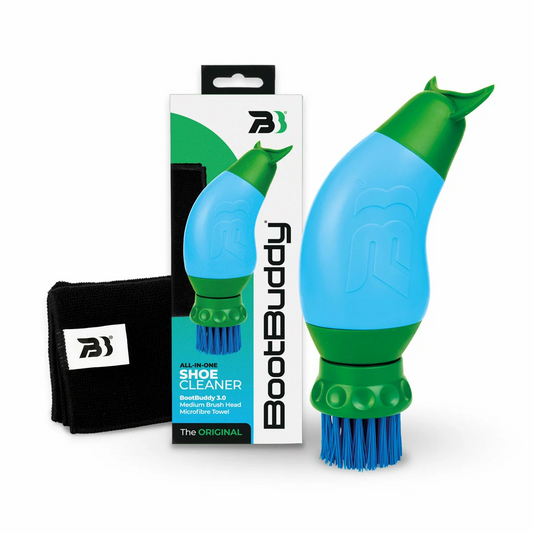 Handheld blue and grey BootBuddy 3-in-1 shoe cleaner scrubbing thick mud off the spiked sole of a football boot outdoors beside a car boot.