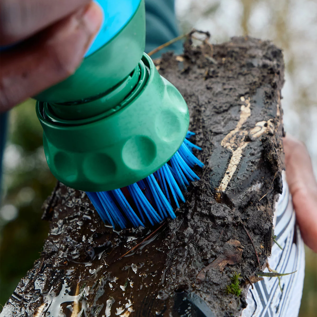 Handheld blue and grey BootBuddy 3-in-1 shoe cleaner scrubbing thick mud off the spiked sole of a football boot outdoors beside a car boot.