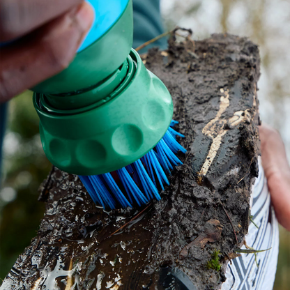 Handheld blue and grey BootBuddy 3-in-1 shoe cleaner scrubbing thick mud off the spiked sole of a football boot outdoors beside a car boot.