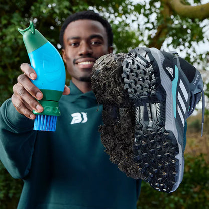 Side angle view of the blue and green BootBuddy shoe cleaner tool resting on a damp patch of grass next to a pair of muddy football boots, highlighting the heavy-duty scraper edge and stiff black bristles.l and consistent flight - 4