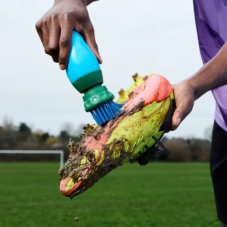 Side angle view of the blue and green BootBuddy shoe cleaner tool resting on a damp patch of grass next to a pair of muddy football boots, highlighting the heavy-duty scraper edge and stiff black bristles.