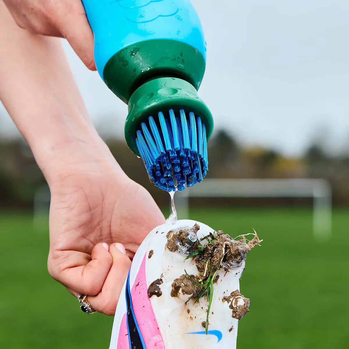 Side angle view of the blue and green BootBuddy shoe cleaner tool resting on a damp patch of grass next to a pair of muddy football boots, highlighting the heavy-duty scraper edge and stiff black bristles.