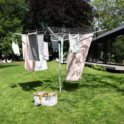 A Brabantia Lift-O-Matic 50m rotary airer standing in a well-maintained garden, featuring taut lines and a protective cover nearby on a clear, sunny day.