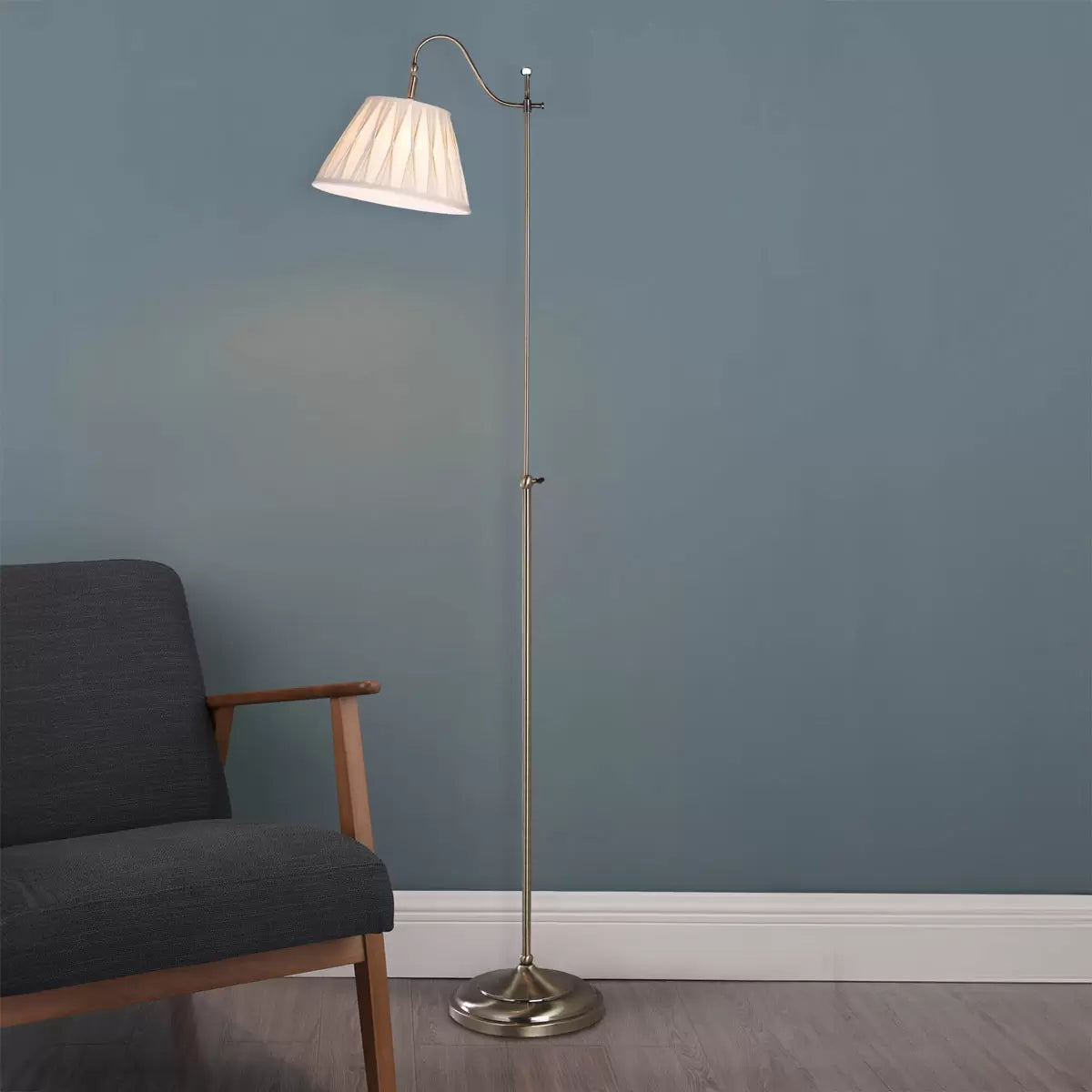 Side view of the Suffolk Rise and Fall Floor Lamp in antique brass, showcasing its adjustable height mechanism and cream pleated faux silk shade next to a classic armchair.