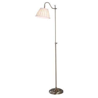 Side view of the Suffolk Rise and Fall Floor Lamp in antique brass, showcasing its adjustable height mechanism and cream pleated faux silk shade next to a classic armchair.