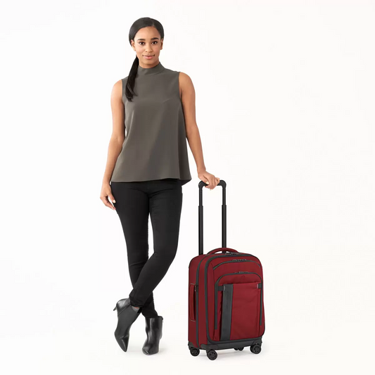Side angle of black Briggs and Riley ZDX 53cm spinner suitcase showing exterior handle rails and durable corner guards in an airport lounge setting.