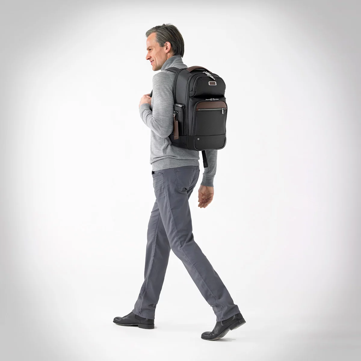Front three-quarter view of the black ballistic nylon Briggs and Riley at work Cargo Backpack standing upright on a minimalist wooden office desk next to a laptop.