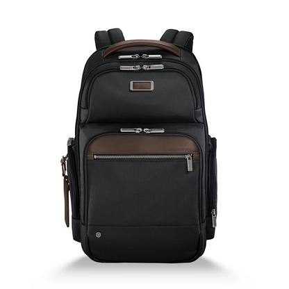 Front three-quarter view of the black ballistic nylon Briggs and Riley at work Cargo Backpack standing upright on a minimalist wooden office desk next to a laptop.
