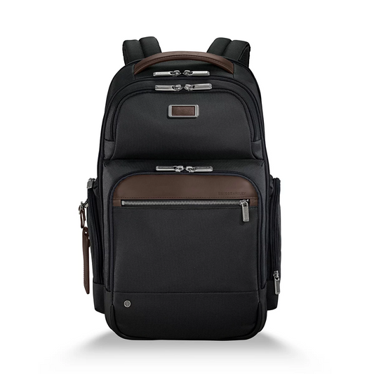 Front three-quarter view of the black ballistic nylon Briggs and Riley at work Cargo Backpack standing upright on a minimalist wooden office desk next to a laptop.