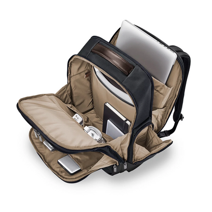 Front three-quarter view of the black ballistic nylon Briggs and Riley at work Cargo Backpack standing upright on a minimalist wooden office desk next to a laptop.