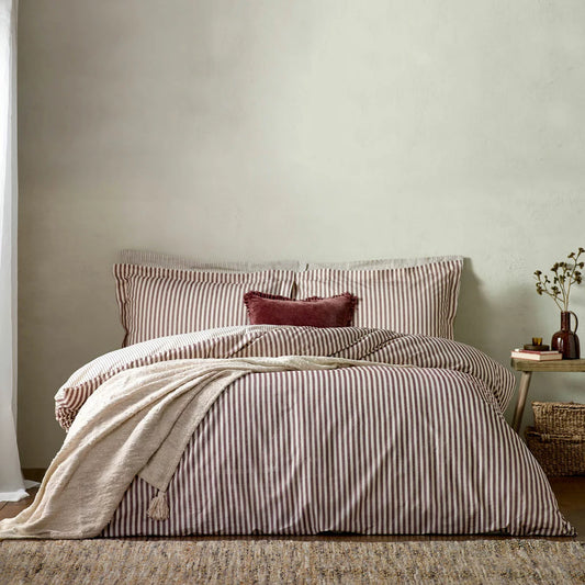 A single bed dressed in the Yard Hebden cotton burgundy striped duvet cover, showcasing the deep red and cream reversible pattern in a warm, cozy bedroom.