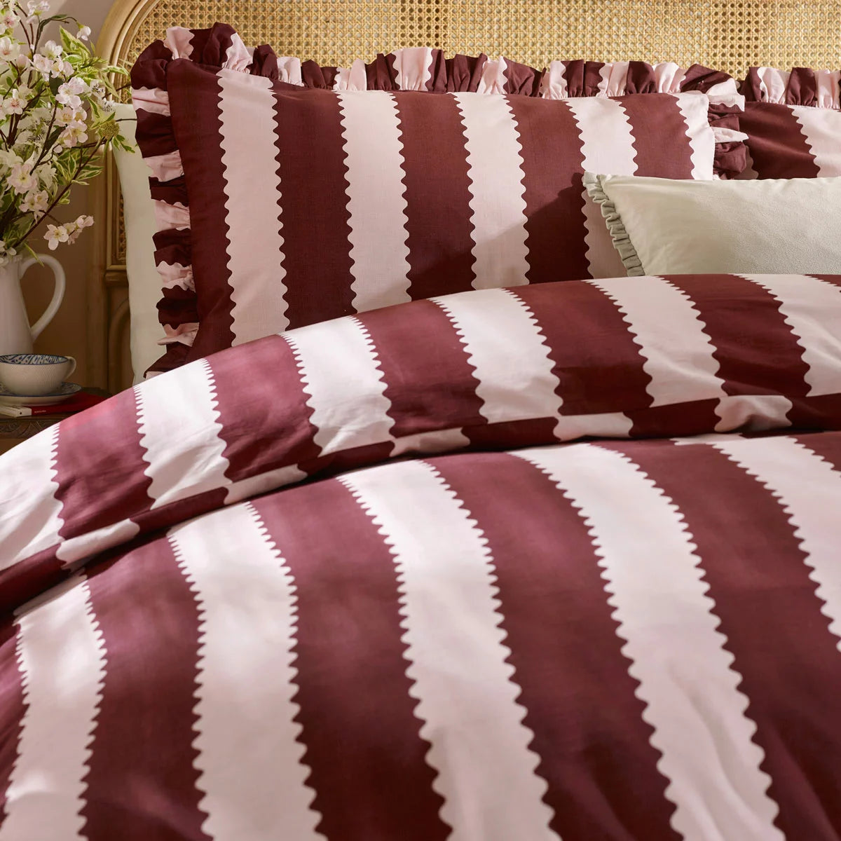 A cozy king bed dressed in the burgundy and white striped Wylder Birdie duvet cover, showing the delicate ruffled edges of the pillowcases resting in a country-style bedroom.