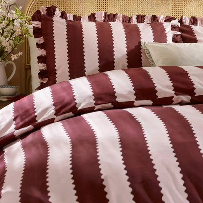 A cozy king bed dressed in the burgundy and white striped Wylder Birdie duvet cover, showing the delicate ruffled edges of the pillowcases resting in a country-style bedroom.