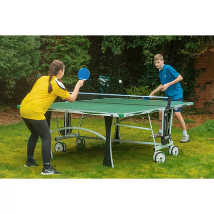 Full side view of the blue Butterfly Premium 4 Outdoor Table Tennis Table fully assembled on a backyard stone patio, showing its silver legs and matching green corner bumpers.
