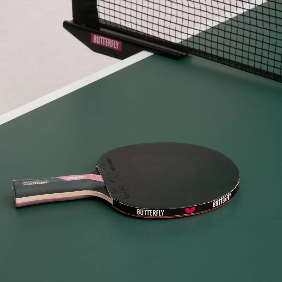 Close-up shot of the red and black Butterfly Timo Boll Ruby table tennis bat showing the anatomic wooden handle and protective edge tape next to three white 40+ training balls on a blue indoor table tennis table.