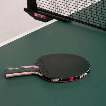 Close-up shot of the red and black Butterfly Timo Boll Ruby table tennis bat showing the anatomic wooden handle and protective edge tape next to three white 40+ training balls on a blue indoor table tennis table.