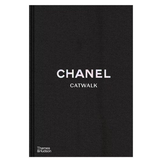 Thick black cloth-bound hardcover book titled Chanel Catwalk with white typography resting on a sleek white coffee table.