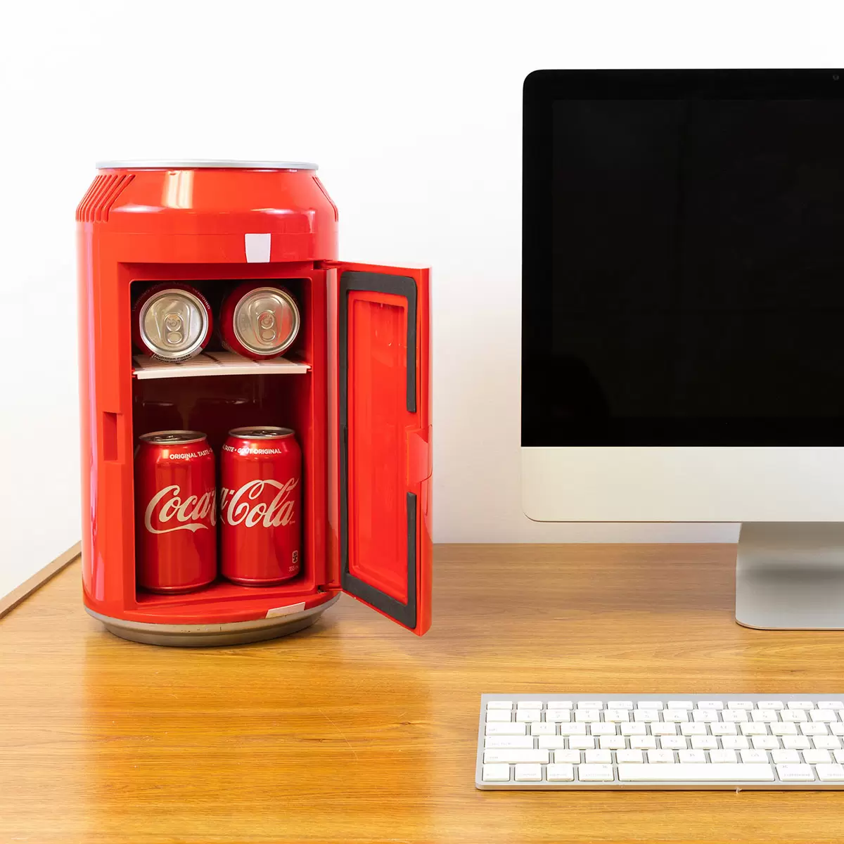 Front view of the vibrant red Coca-Cola can-shaped mini fridge with silver pull-tab handle, standing on a white minimalist office desk next to a laptop.