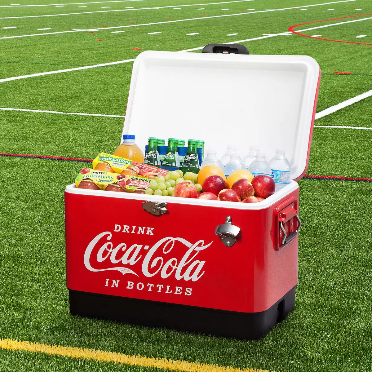 Red vintage style Coca-Cola 51L stainless steel ice chest cooler with built-in bottle opener standing on a rustic wooden patio table during an outdoor gathering.