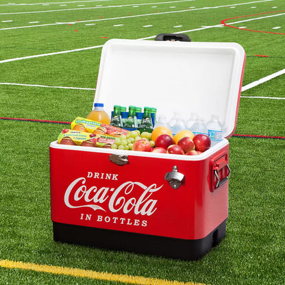 Red vintage style Coca-Cola 51L stainless steel ice chest cooler with built-in bottle opener standing on a rustic wooden patio table during an outdoor gathering.