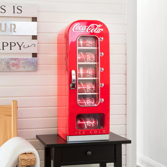 Front view of the vibrant red Coca Cola 5L mini fridge vending machine featuring a clear vertical display window full of cans, polished chrome accents, and retro artwork on a wooden desk.
