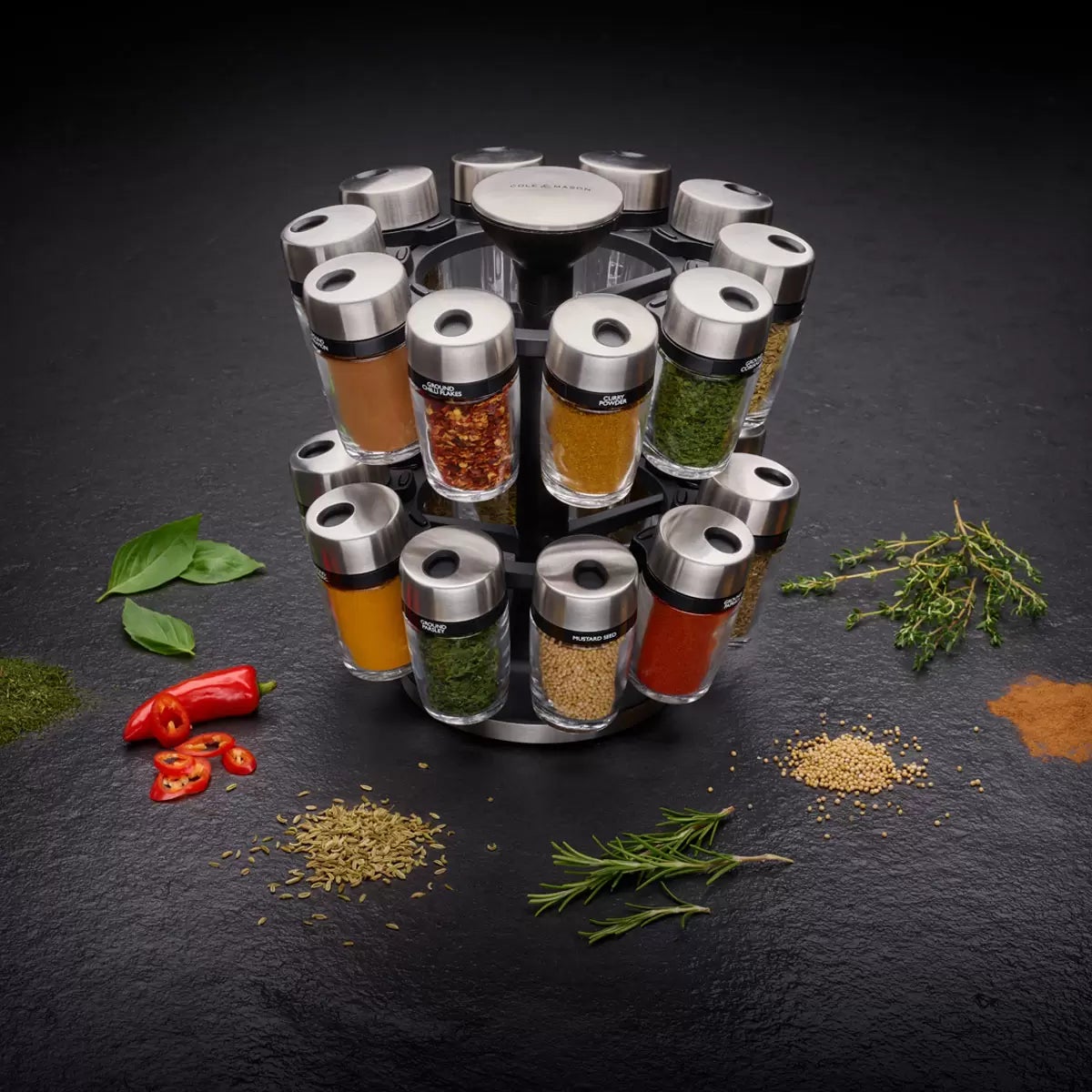 Stainless steel Cole & Mason 20 jar spice carousel filled with colorful herbs and spices sitting on a clean granite kitchen countertop.