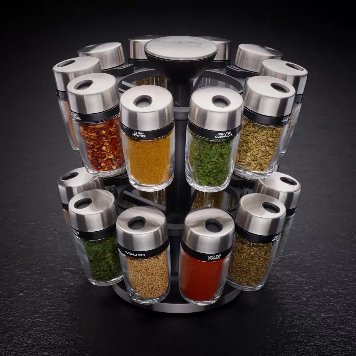 Stainless steel Cole & Mason 20 jar spice carousel filled with colorful herbs and spices sitting on a clean granite kitchen countertop.