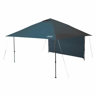 A large blue 13 by 13 foot Coleman Oasis pop-up canopy with an attached white fabric side shade wall standing securely on a green grassy lawn with guy lines pinned to the earth under a bright blue sky.