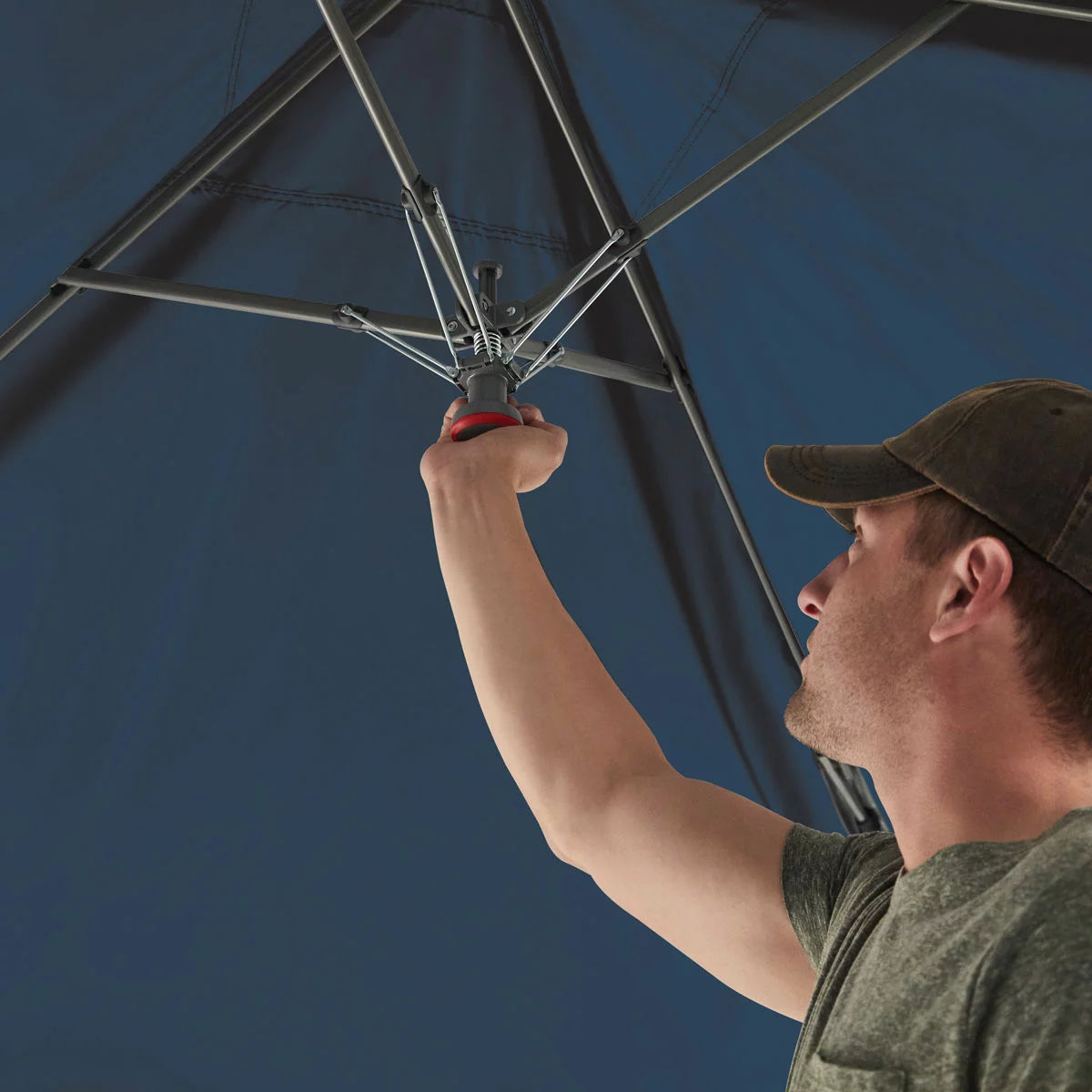 A large blue 13 by 13 foot Coleman Oasis pop-up canopy with an attached white fabric side shade wall standing securely on a green grassy lawn with guy lines pinned to the earth under a bright blue sky.