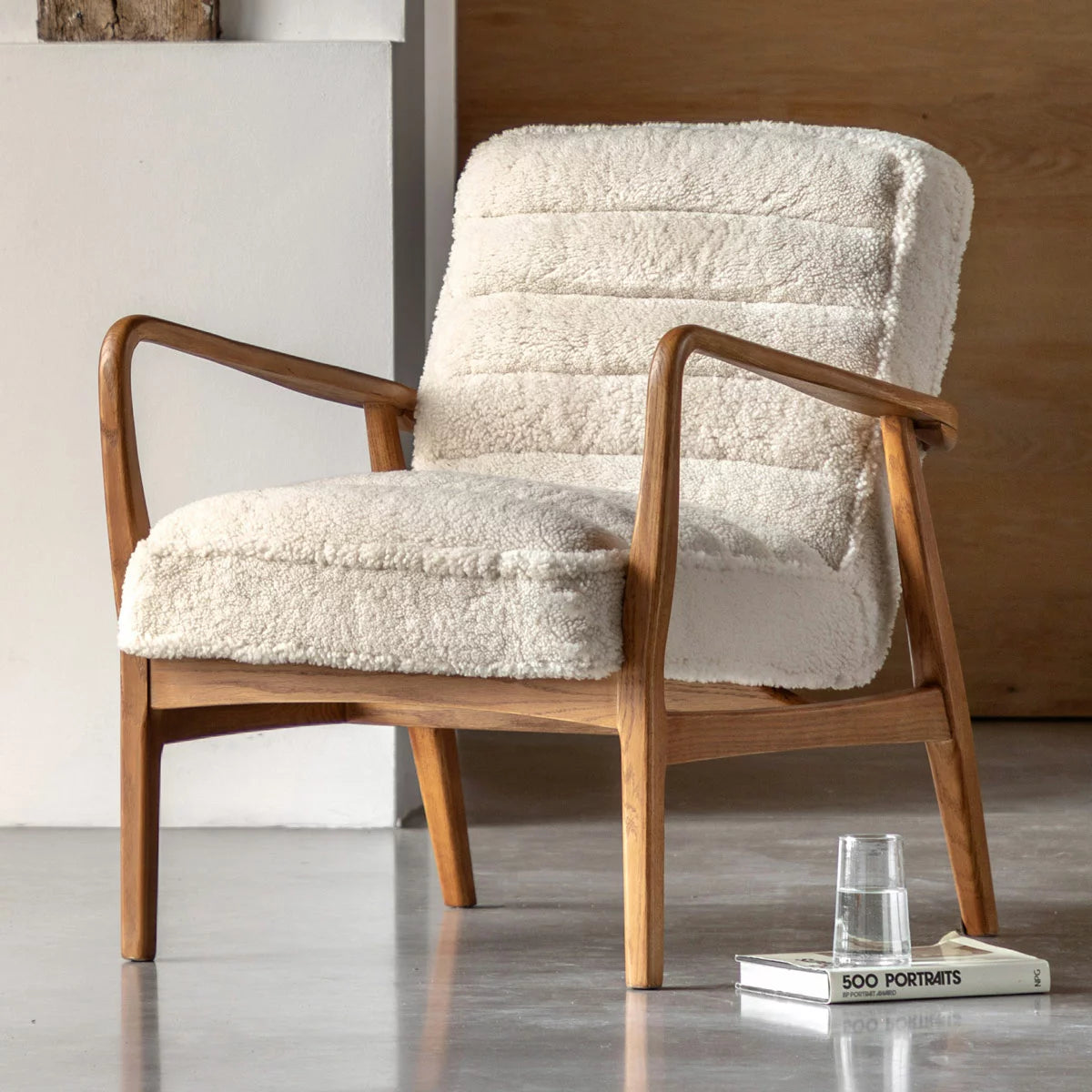A three-quarter view of the Gallery Datsun Sheepskin Fabric Armchair showcasing its soft cream upholstery, curved wooden armrests, and natural ash wood frame in a bright living room corner.